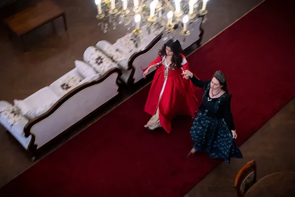 Vista cenital de dos mujeres con vestidos de fantasía caminando sobre una alfombra roja; a la derecha se aprecia Forest en verde oscuro con corona.