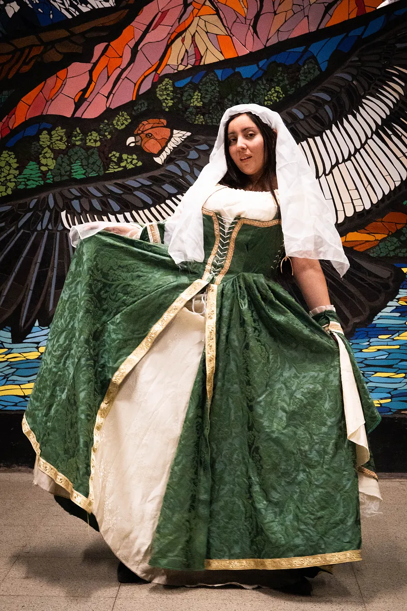 Vestido verde con velo y corsetería dorada frente a un vitral, una lectura élfica y ceremonial de la fantasía barroca.
