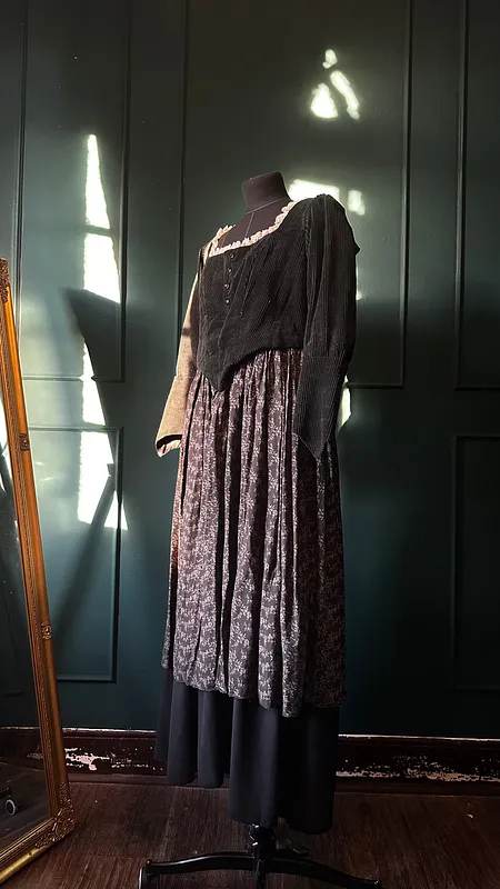 Vestido Katrin sobre maniquí en vista lateral, con cuerpo negro texturado, manga larga amplia, falda floral fruncida y capa inferior negra.