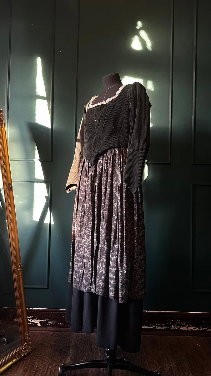 Vestido Katrin sobre maniquí en vista lateral, con cuerpo negro texturado, manga larga amplia, falda floral fruncida y capa inferior negra.
