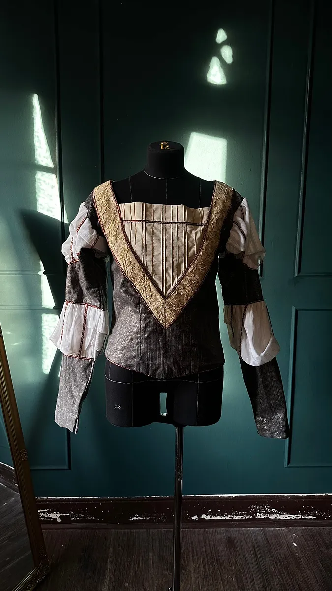Vista frontal del jubón sobre maniquí, con cuerpo gris texturado, cuello amplio dorado y mangas blancas divididas por bandas oscuras.