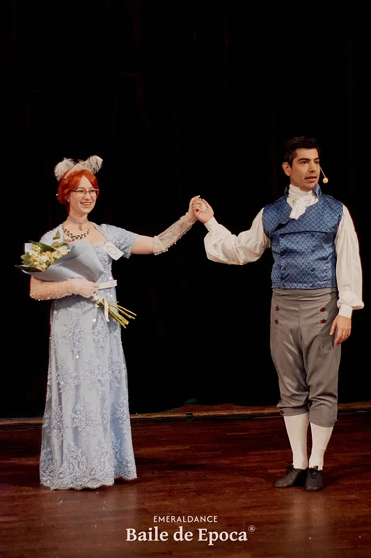 Pareja sobre un escenario; ella lleva vestido celeste de encaje y sostiene flores, mientras él viste chaleco azul y pantalón corto.