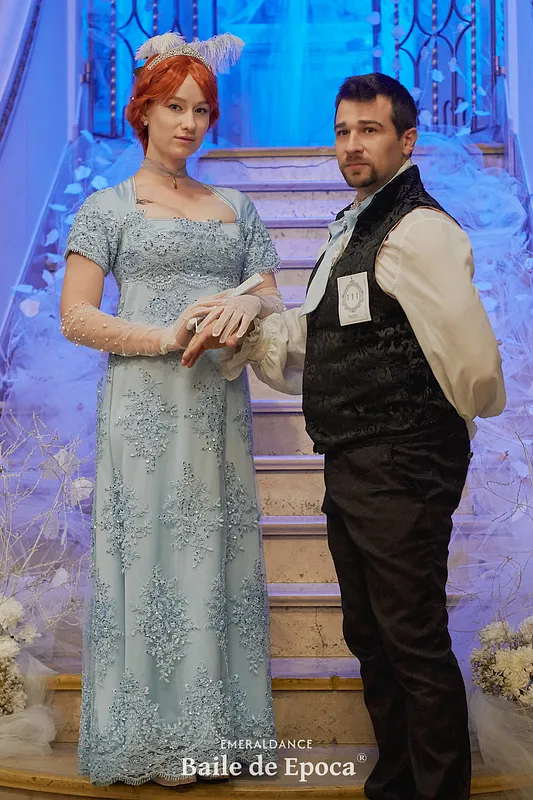 Retrato de pareja ante una escalera, con el vestido celeste bordado mostrado de cuerpo completo junto a un conjunto masculino oscuro.