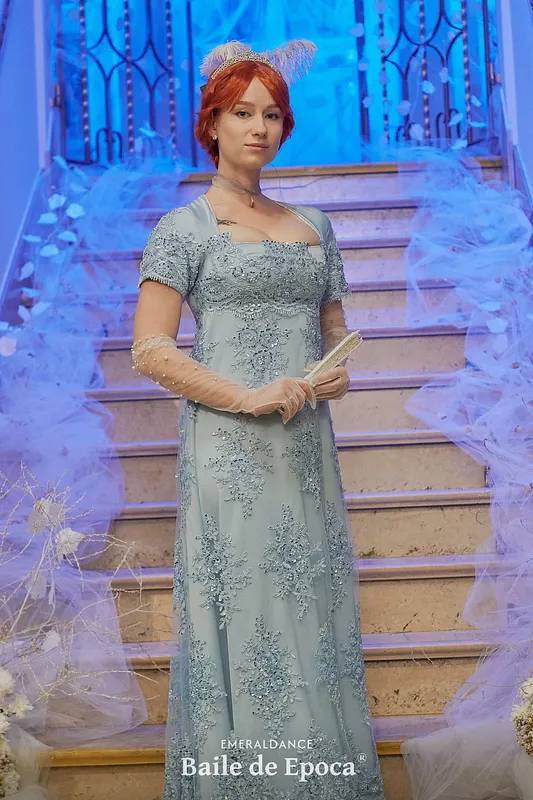 Mujer frente a una escalera con vestido celeste largo bordado, mangas cortas, guantes transparentes y abanico sostenido entre las manos.