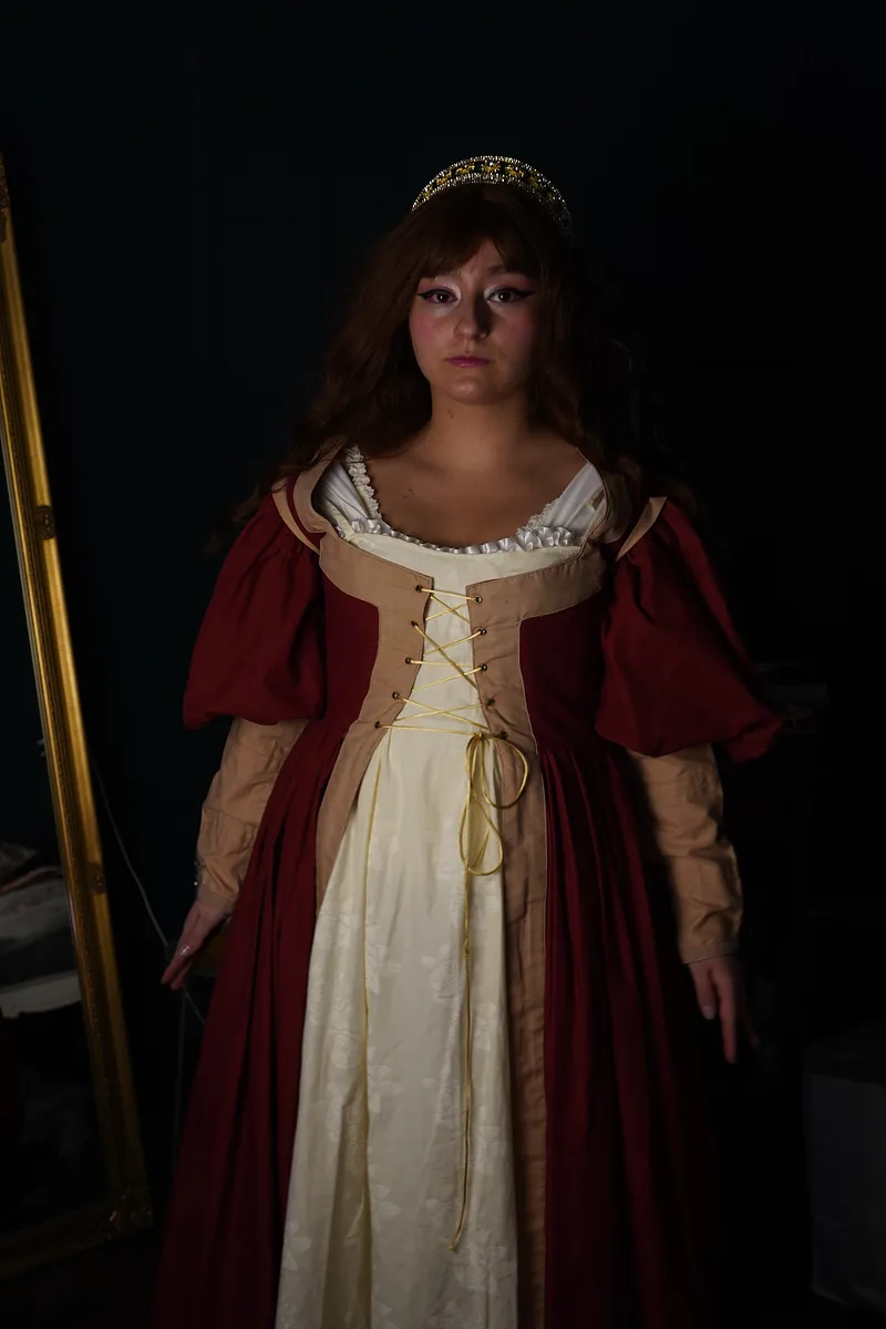 Vista frontal del vestido rojo medieval con panel crema, cordones dorados, mangas abullonadas y tocado tipo tiara sobre cabello largo.