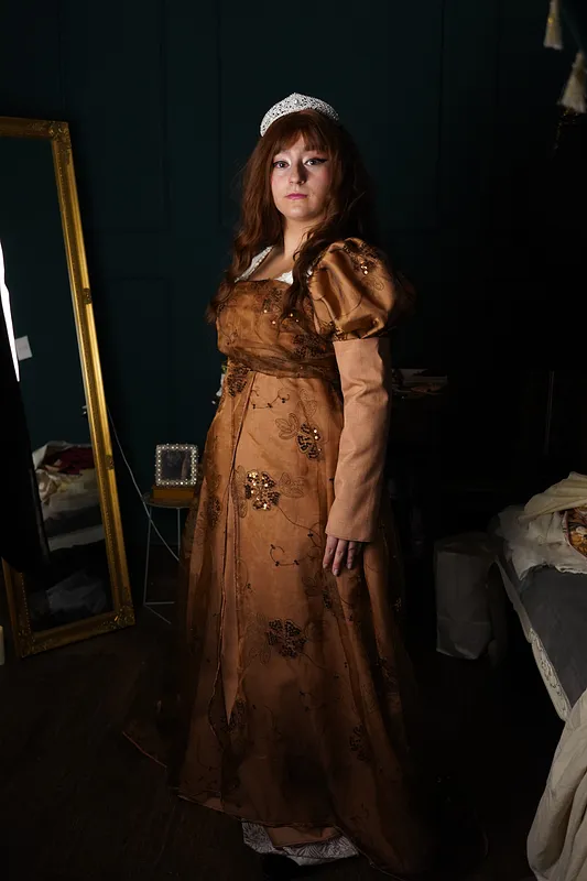 Vista de cuerpo completo del vestido Lilian en una modelo con tiara, mostrando la falda larga cobriza, mangas abullonadas y sobrefalda bordada en un interior oscuro.