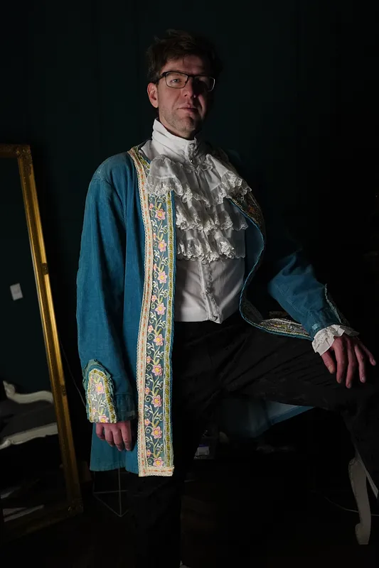 Hombre sentado con abrigo largo verde azulado, pechera clara bordada, cuello blanco con encaje y puños ornamentados.