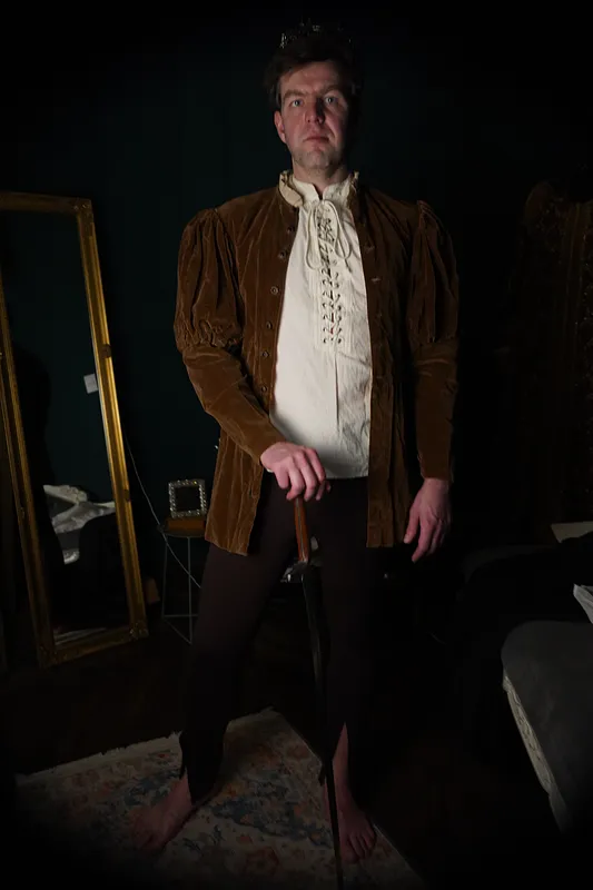 Hombre de frente con chaqueta de terciopelo ocre, hombros fruncidos, camisa marfil entrelazada y calzas oscuras.