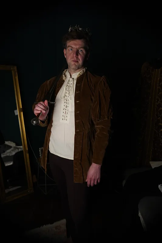 Retrato frontal del conjunto ocre con camisa marfil entrelazada, mientras el hombre sostiene el pomo de una espada.