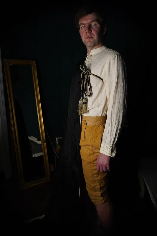 Hombre en vista tres cuartos con capa negra abierta, camisa marfil de cuello alto y pantalones mostaza hasta la rodilla.