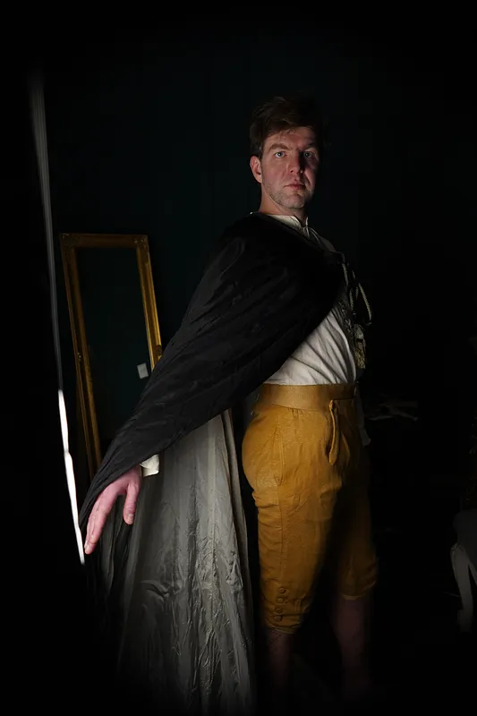 Hombre de pie con capa negra drapeada sobre los hombros, camisa clara de lazo y pantalones cortos color mostaza.
