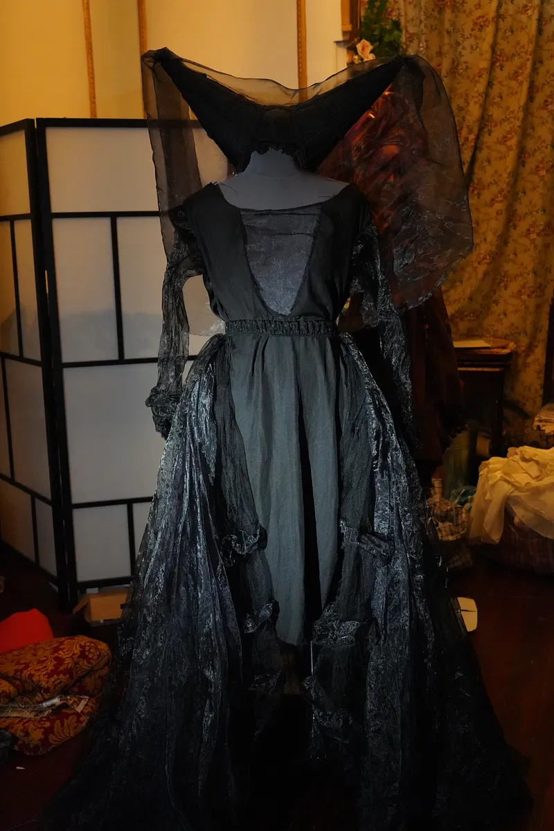 Vista posterior del vestido sobre maniquí, con espalda negra, cintura recogida, mangas oscuras y gran sobrefalda brillante en capas; el velo se abre hacia ambos lados.