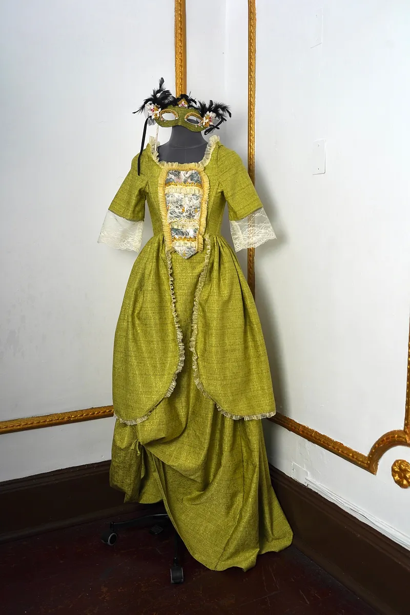 Vista frontal del vestido verde oliva sobre maniquí, con antifaz decorado, escote con vuelos claros, panel central de encaje y sobrefalda amplia con ribete fruncido.