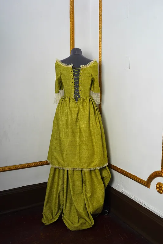 Vista trasera del vestido sobre maniquí, con espalda abierta ajustada por cordones cruzados, vuelos en el escote, falda amplia y bajo con franja clara fruncida.