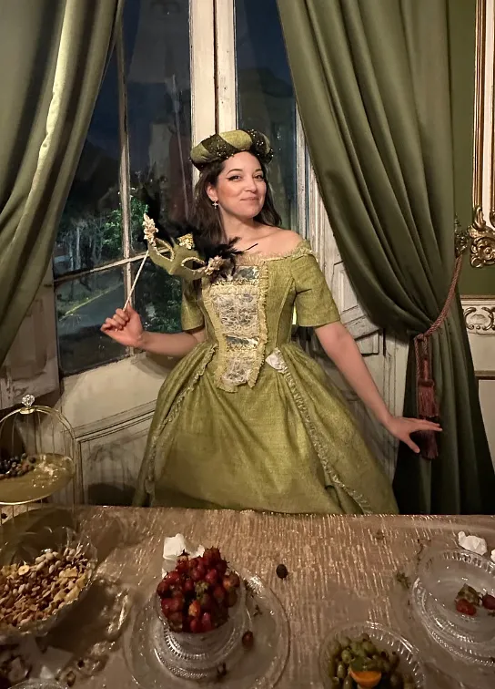 Persona usando el vestido verde en un interior con cortinas, sosteniendo un antifaz de varilla; se ven el tocado a juego, el corpiño decorado y la falda voluminosa.
