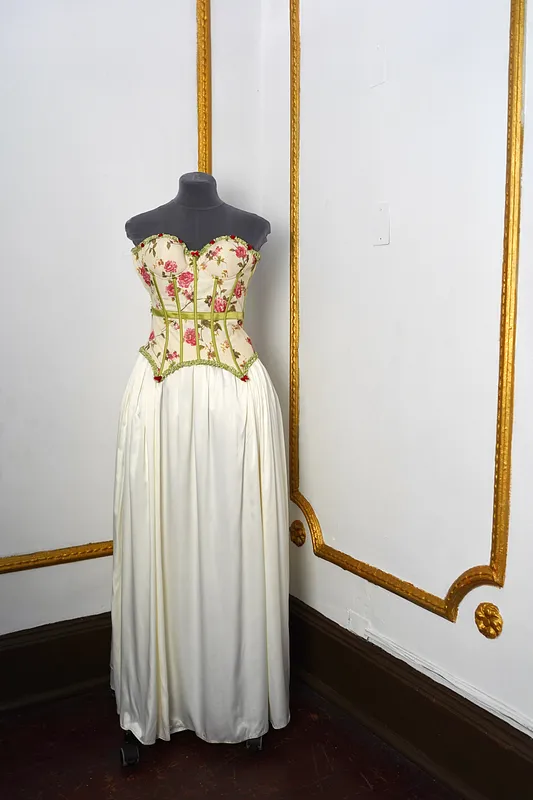 Vista frontal de vestido largo en maniquí, con corset floral strapless, varillas verdes, ribetes decorativos y falda marfil amplia.