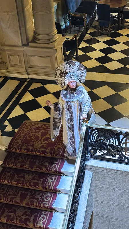 Persona usando el traje en una escalera con alfombra roja, mostrando la caída larga del vestido, el tocado alto y una manga extendida.