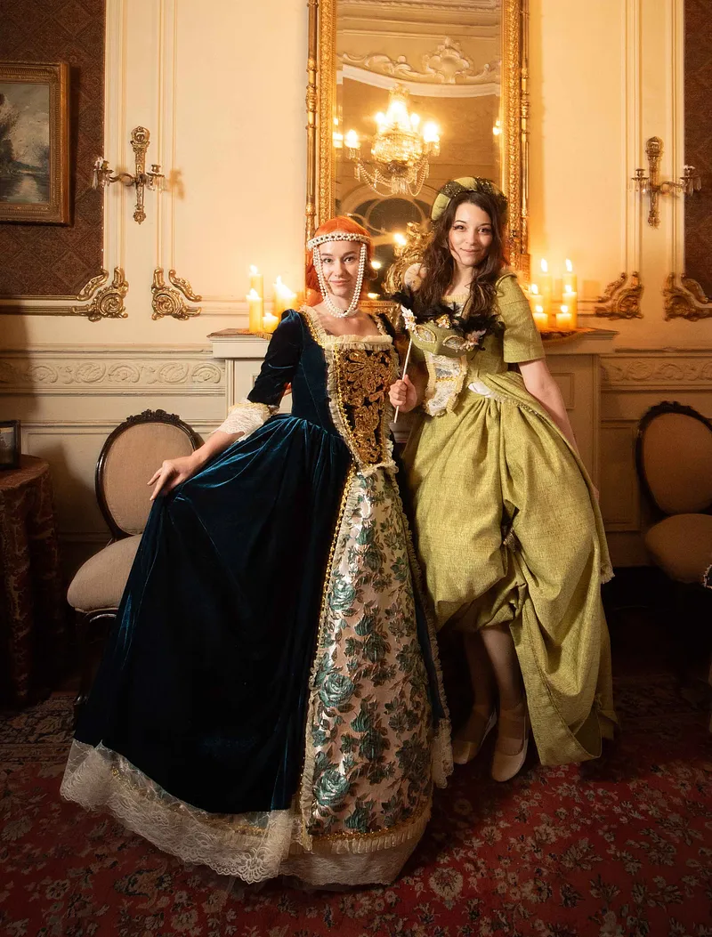 Dos personas en un salón antiguo; una lleva el vestido azul con panel floral y bordado dorado, junto a otra con vestido verde claro.
