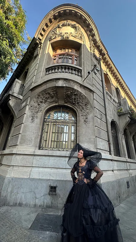 Persona posa en la calle con vestido negro gótico de falda amplia, cuerpo decorado con motivo dorado, mangas transparentes y velo estructurado frente a una fachada antigua.