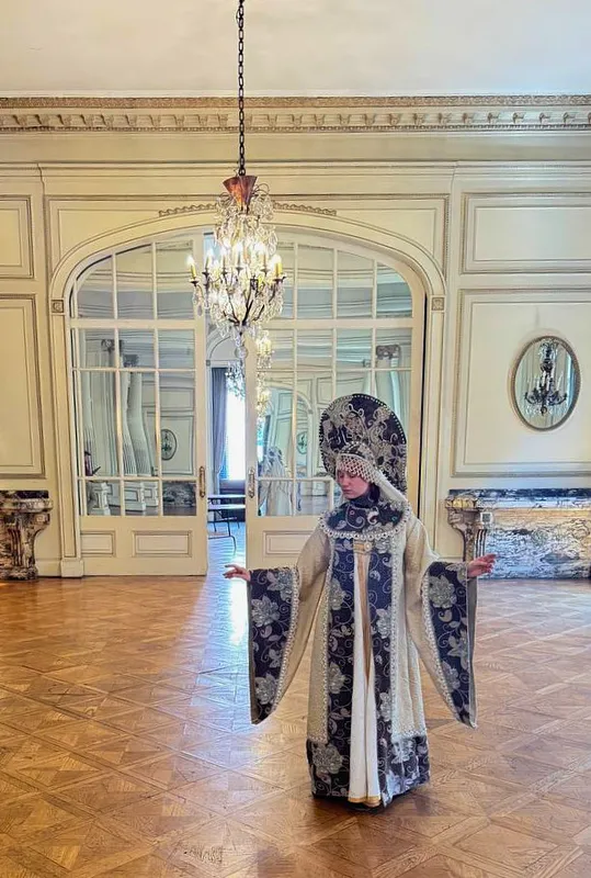 Vista de cuerpo completo en un salón elegante, con el vestido ceremonial abierto hacia los costados y el tocado destacado bajo una lámpara.