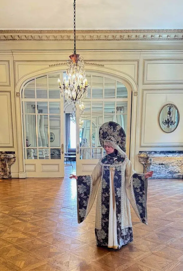 Vista de cuerpo completo en un salón elegante, con el vestido ceremonial abierto hacia los costados y el tocado destacado bajo una lámpara.