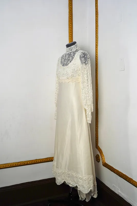 Vista frontal lateral de vestido largo marfil en maniquí, con cuello alto de encaje, mangas largas transparentes, cuerpo bordado y falda fluida.