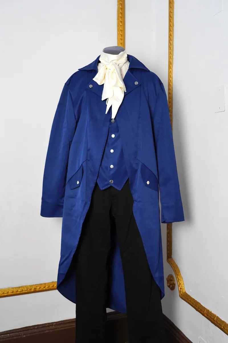 Vista frontal del conjunto en maniquí, con levita azul larga abierta, chaleco azul abotonado, pañuelo crema al cuello y pantalón negro.