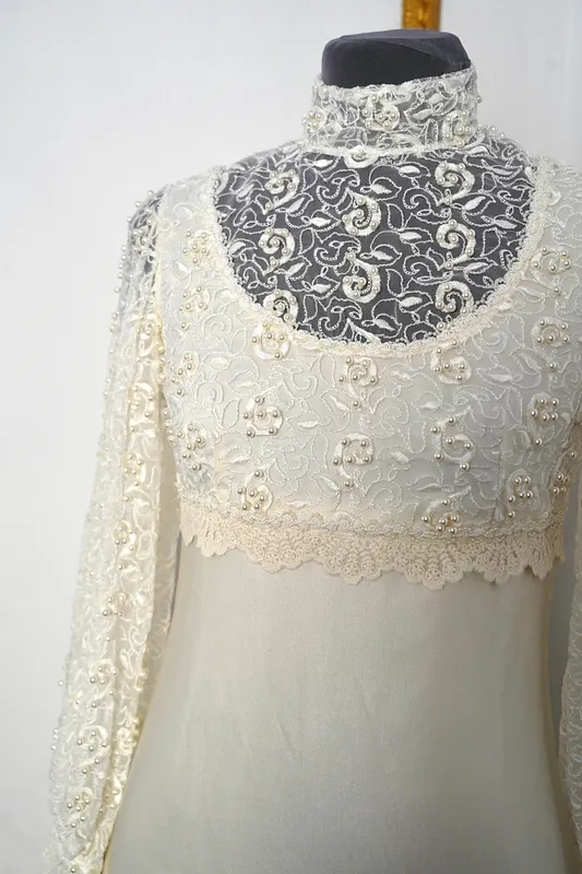 Detalle frontal del cuerpo del vestido, con cuello alto, canesú de encaje, escote amplio, bordados florales y perlitas sobre tela marfil.
