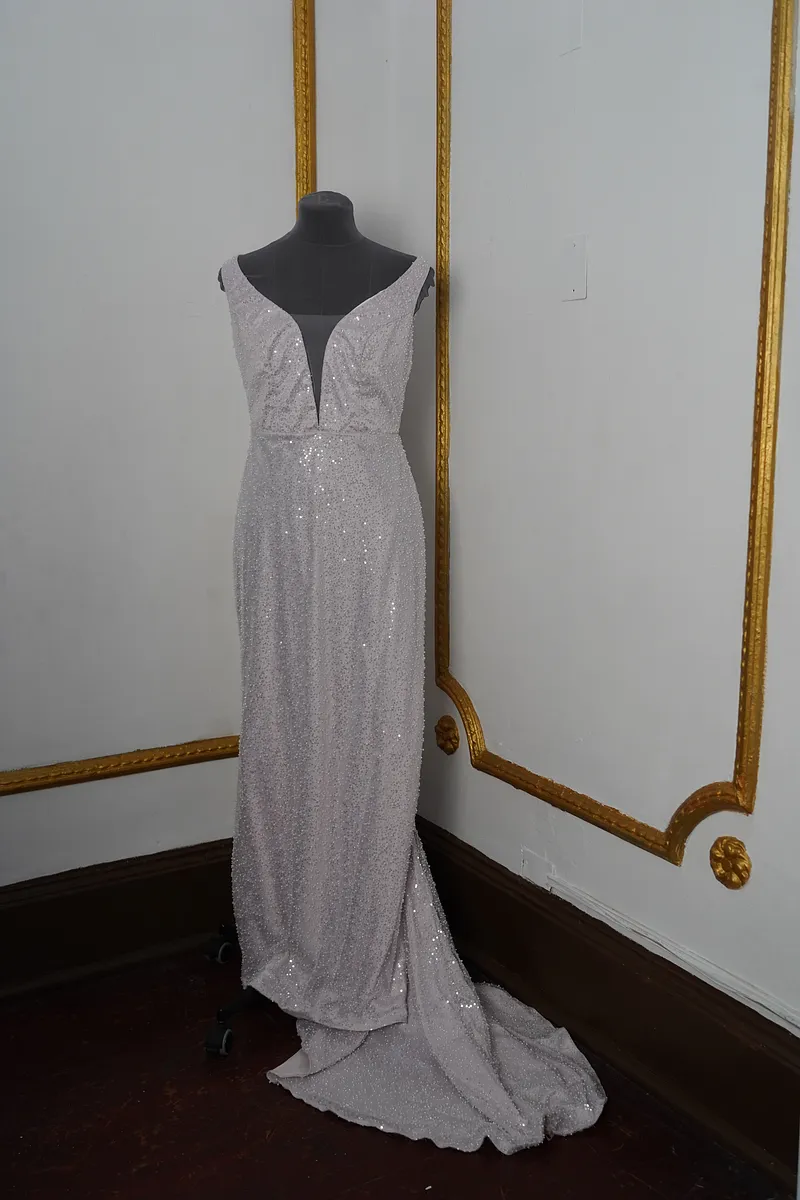 Vista frontal completa del vestido en maniquí, con tirantes anchos, escote profundo, falda larga plateada brillante y cola recogida hacia un costado.