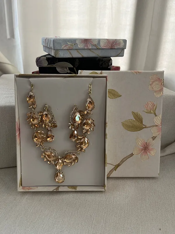 Collar dorado de diseño floral con cristales, cuentas tipo perla y colgante en forma de lágrima, presentado dentro de una caja decorada.