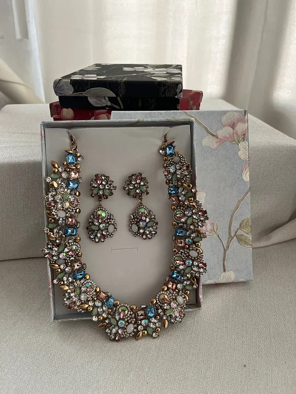 Collar ancho y aros a juego, formados por piedras brillantes multicolores con monturas metálicas, presentados sobre una caja clara.