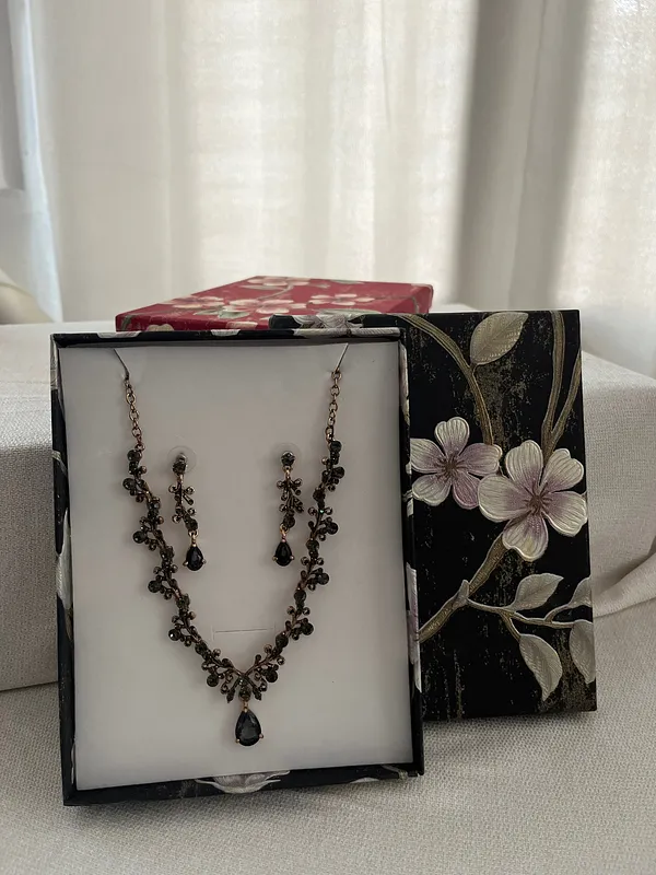 Conjunto de collar y aros con piedras oscuras y pequeños detalles metálicos, dispuesto dentro de una caja blanca de presentación.