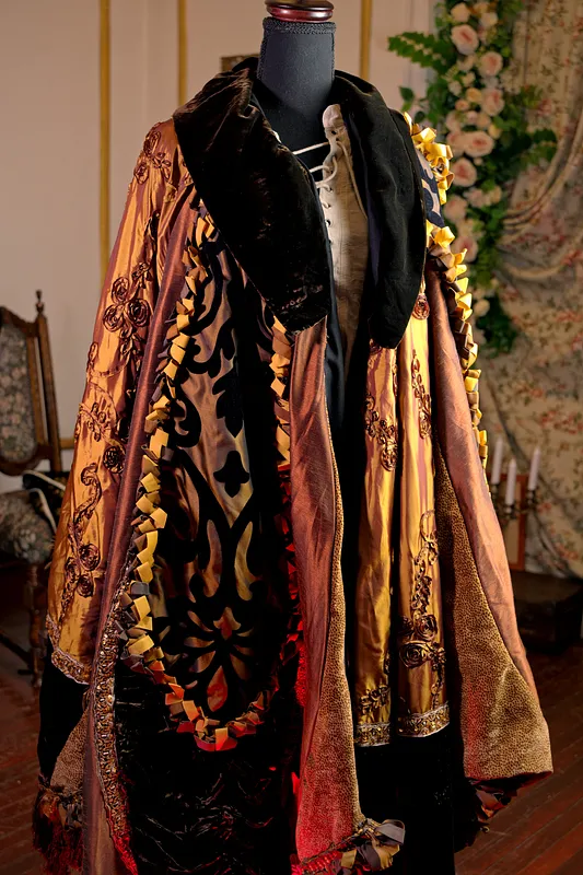 Capa amplia de brocado dorado y negro sobre maniquí, con cuello de piel oscura y destellos del forro rojo interior.