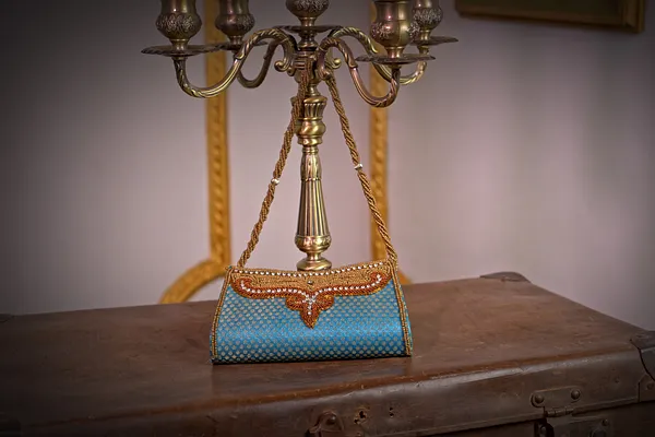Cartera de fiesta turquesa con superficie de cuentas, detalle frontal anaranjado y dorado, y cadena metálica larga.