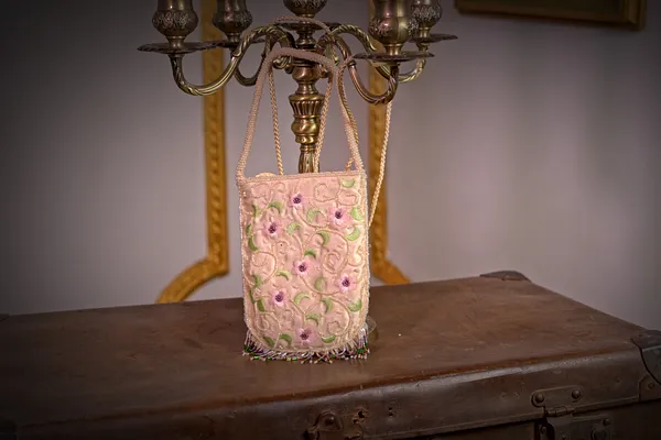 Bolso rectangular rosado con correa delgada, superficie bordada con pequeñas flores y aplicaciones brillantes, colgado frente a un muro claro.