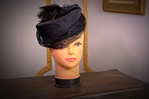 Sombrero negro pequeño sobre cabeza de maniquí, con copa baja, ala inclinada, cintas superpuestas y un adorno de plumas.