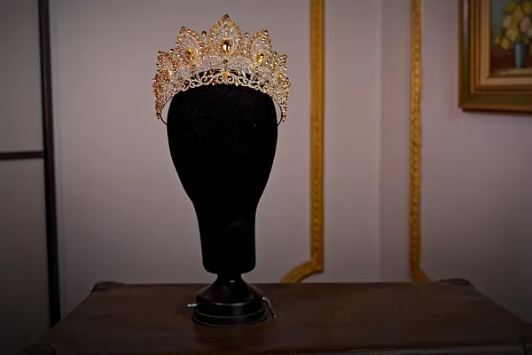Corona dorada abierta sobre soporte negro, con puntas ornamentadas, filigrana y numerosas piedras transparentes de distintos tamaños.