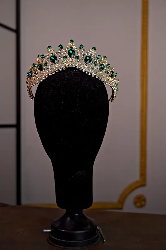 Tiara plateada sobre soporte negro, decorada con piedras verdes ovaladas, cristales transparentes y pequeñas puntas ornamentales.