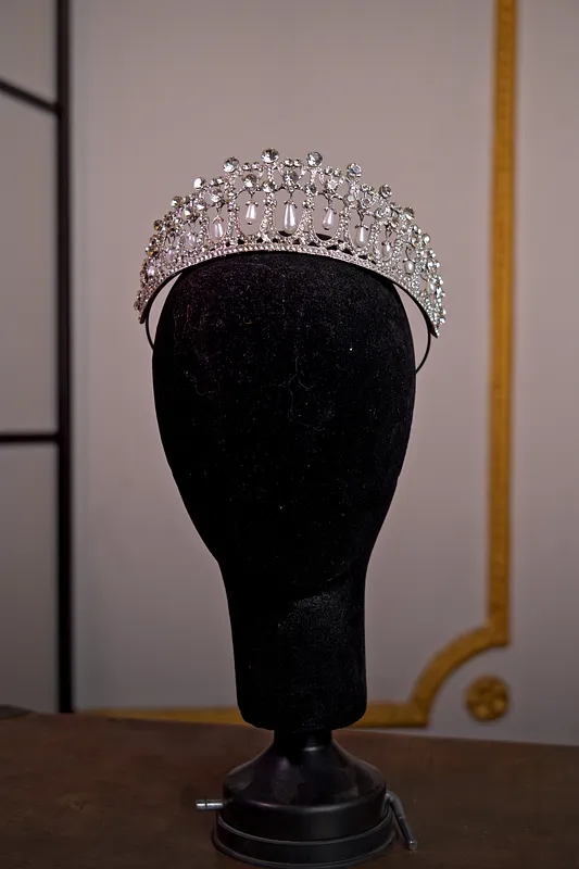 Tiara plateada de cristales transparentes sobre cabeza de maniquí, con arcos simétricos y una hilera superior de pequeñas piedras redondas.