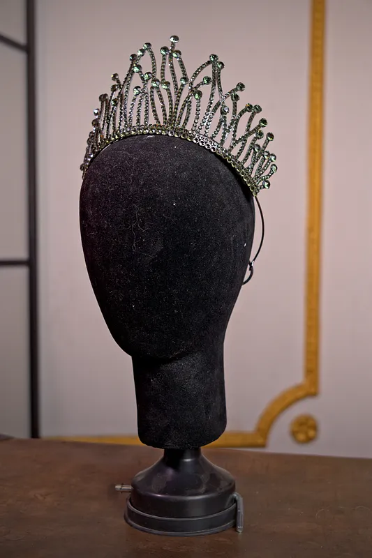 Tiara plateada alta con estructura de puntas curvas y pequeñas piedras verdes, presentada sobre una cabeza de maniquí negra.