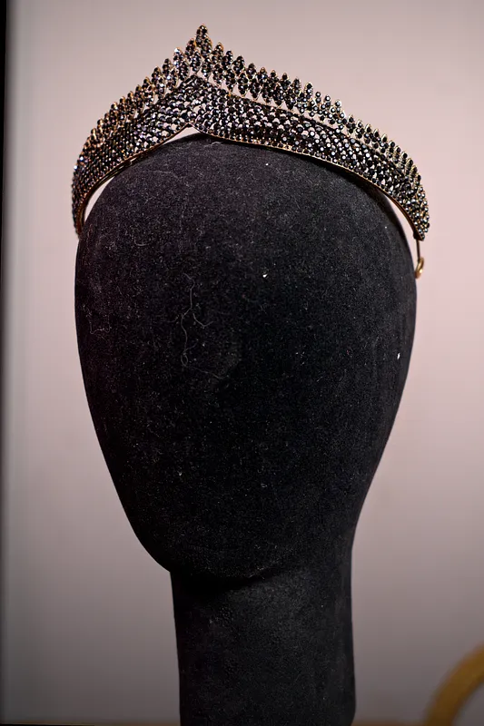 Tiara alta de metal oscuro con diseño calado y numerosas puntas, cubierta por pequeñas piedras brillantes en varias hileras.