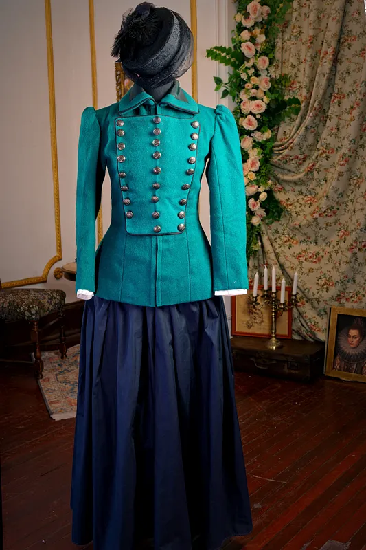 Vista frontal del conjunto con chaqueta turquesa de doble botonadura, falda larga azul marino y pequeño sombrero negro.