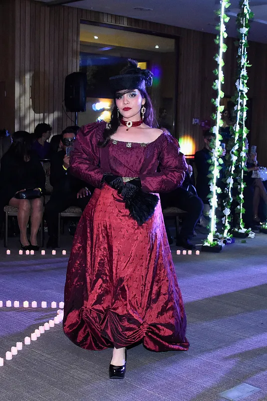 Modelo usando el vestido burdeo completo en pasarela, con escote amplio, mangas abullonadas, falda recogida al frente, sombrero negro, guantes y accesorios oscuros.