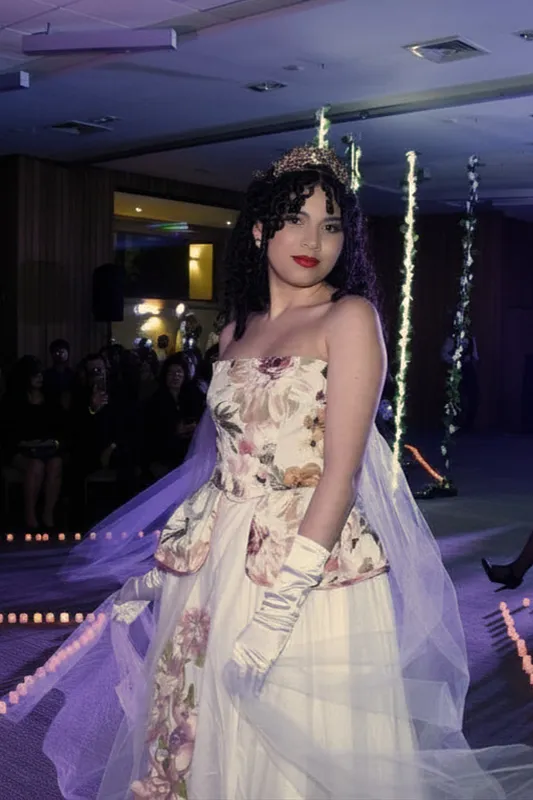 Modelo en pasarela interior con vestido strapless floral, sobrefalda peplum, falda blanca con capas de tul translúcido y guantes claros de estilismo.