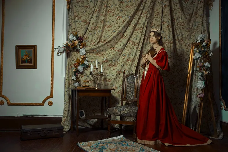 Vestido rojo medieval en escenario palaciego, con falda larga extendida, libro en las manos, espejo dorado y cortinaje floral.