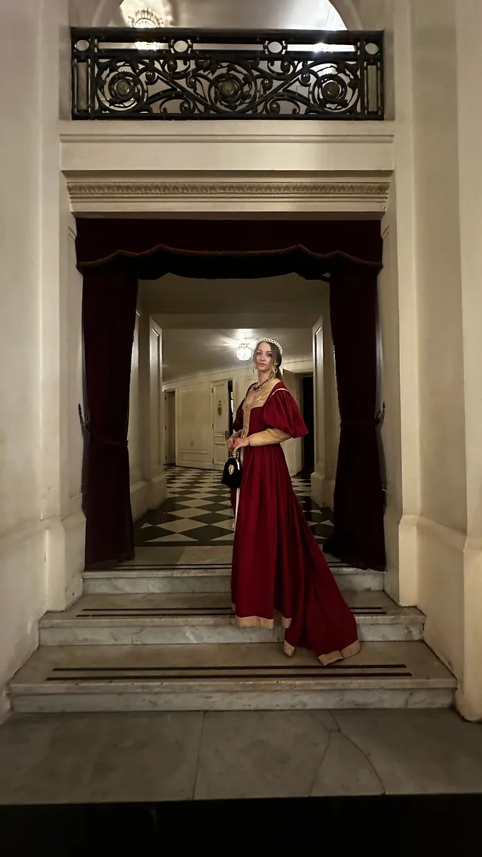 Vestido rojo largo usado en un acceso con mármol, cortinas granate y piso ajedrezado, combinado con tocado y bolso pequeño.