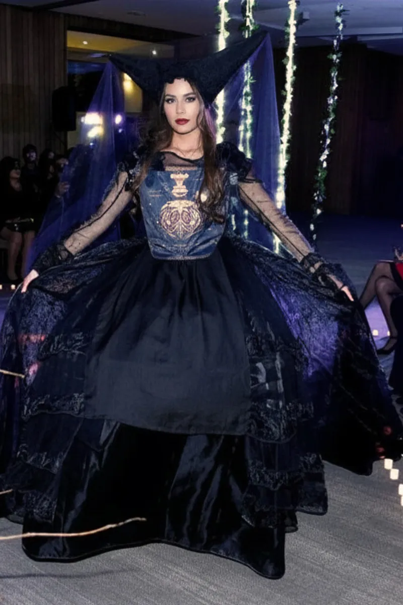 Modelo en pasarela muestra el vestido de frente con falda negra amplia, sobrefalda brillante extendida, mangas de tul y tocado triangular con velo translúcido.