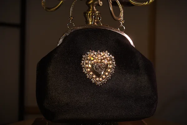 Pequeño bolso negro de tela con cierre metálico, asa corta y broche central en forma de corazón rodeado de pedrería.