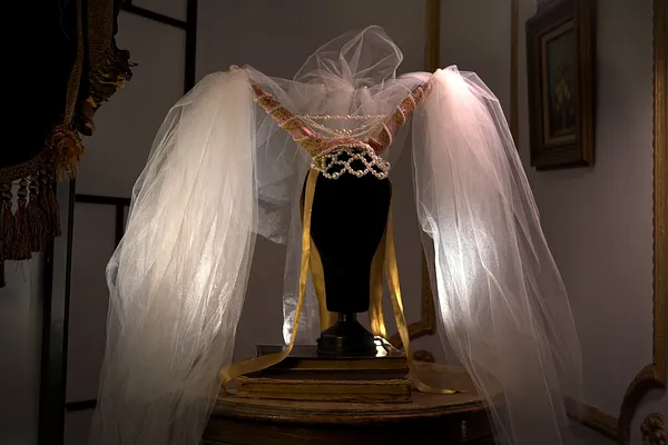 Tocado de fantasía sobre maniquí, con grandes capas blancas de tul y plumas extendidas, sujetas por cintas doradas y adorno central.