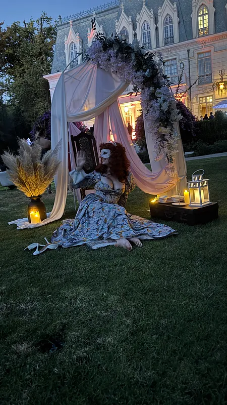 Modelo sentada sobre el pasto con el vestido floral extendido, antifaz blanco y montaje exterior con telas, flores, faroles y fachada iluminada al atardecer.